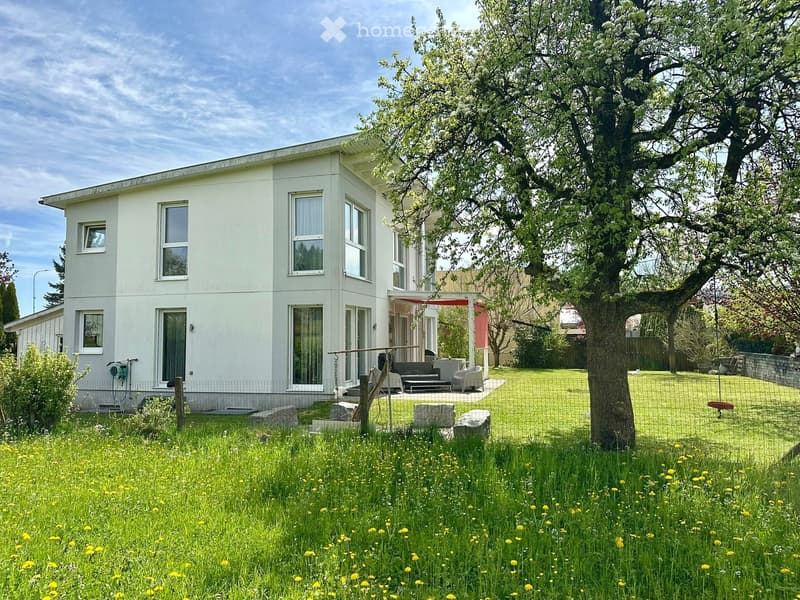 Single-story white house with large windows, surrounded by a well-maintained garden with a swing, seating area, and a large tree.