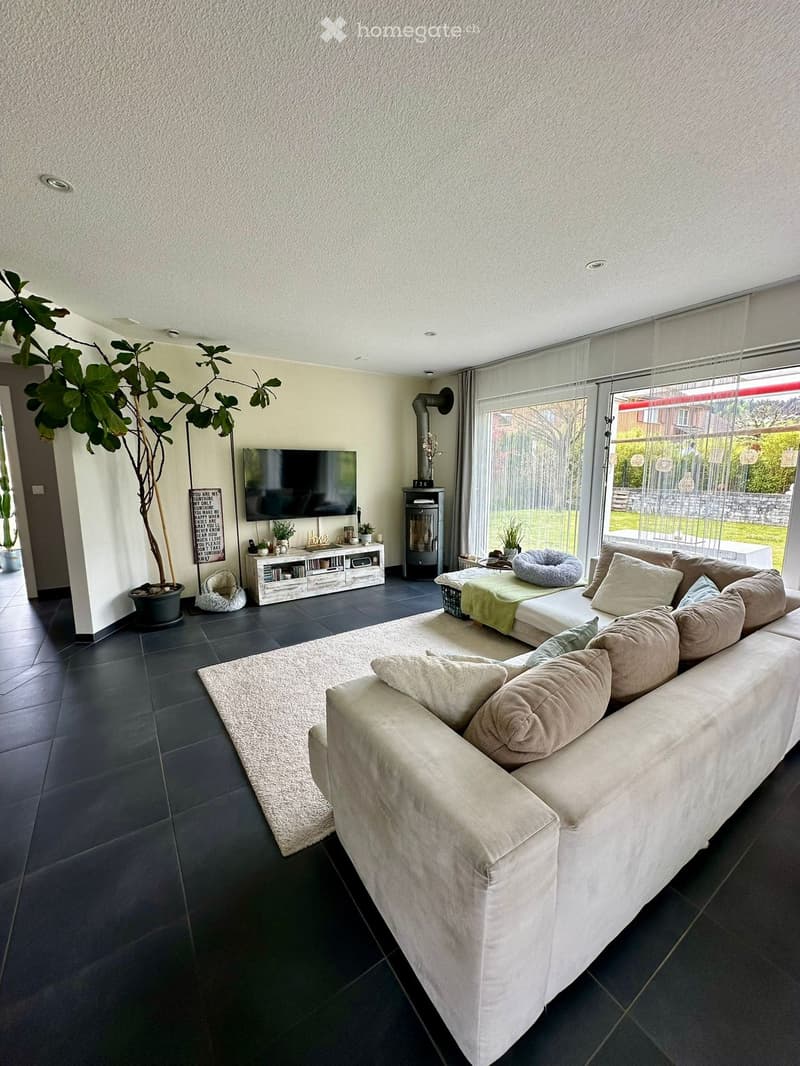 Modern living room with a large sectional sofa, coffee table, television, fireplace, and large windows with a view of the garden.