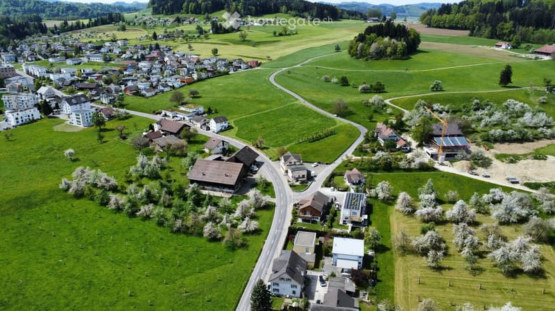 Aerial view of a rural village with scattered houses, green fields, and winding roads surrounded by hills and forests.