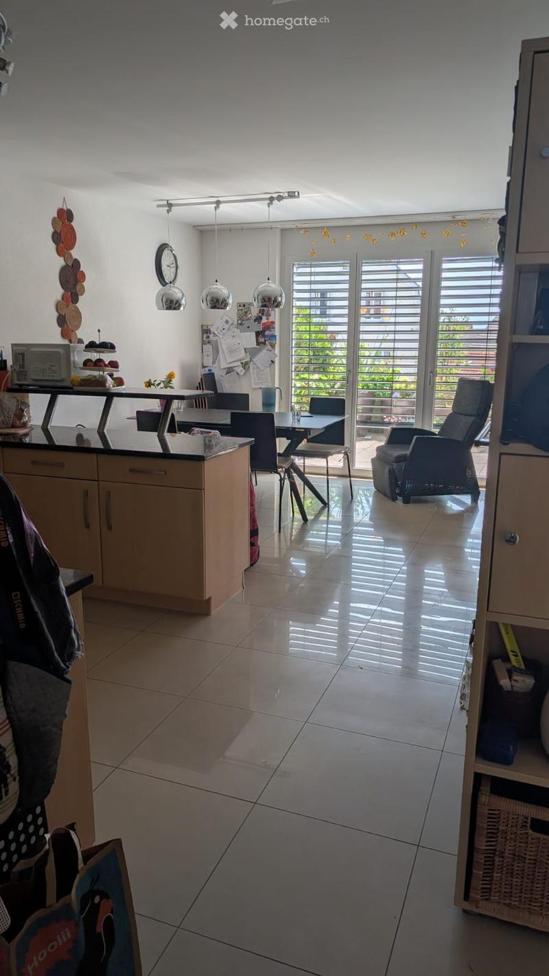 Modern kitchen with a central island, pendant lights, and a dining area with chairs and a table. The space features a tiled floor and large windows with blinds, offering a view of the outdoor area.