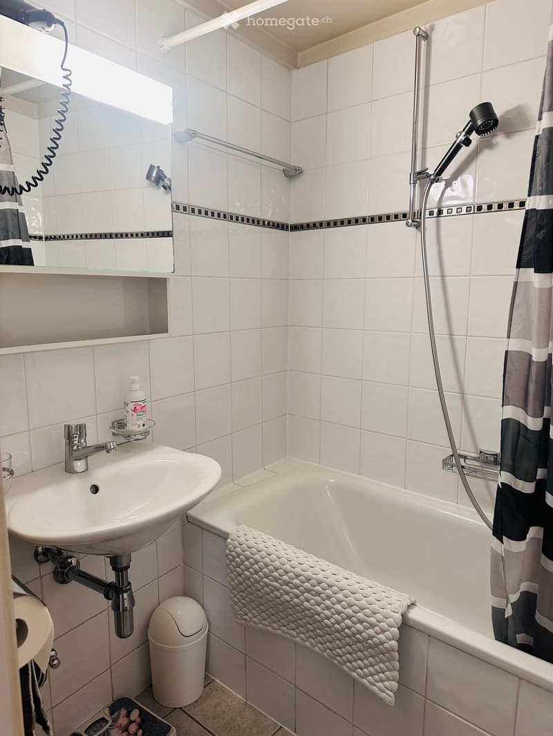 A small bathroom with a white tiled wall, a sink with a mirror above it, a bathtub with a towel draped over the edge, a shower curtain, and a toilet. A bottle of shampoo is placed on the sink.