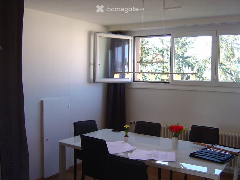 Bright room with large windows, a white table surrounded by black chairs, two vases with flowers, and papers on the table.