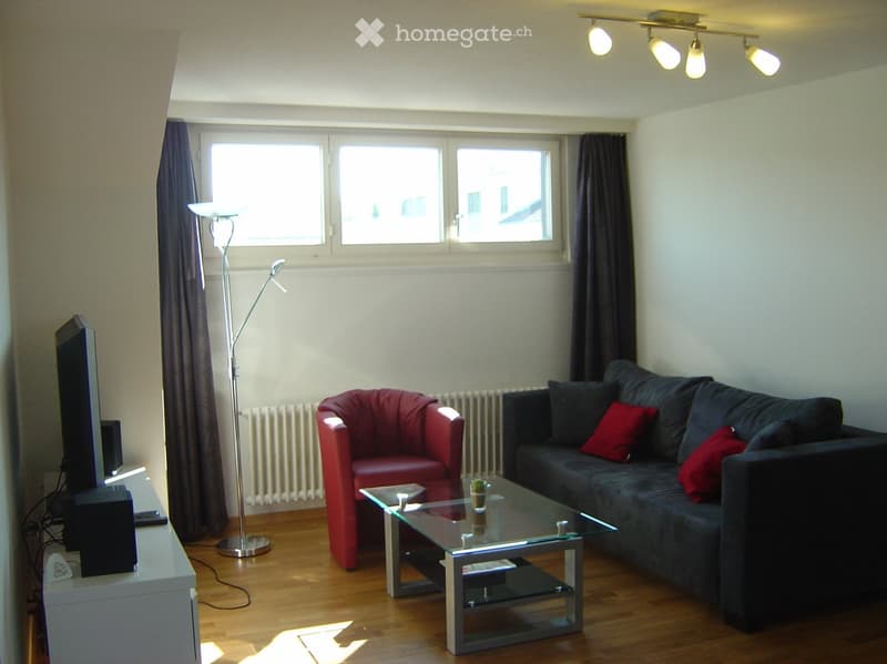 Modern living room with wooden flooring, large windows, grey sofa with red cushions, red armchair, glass coffee table, TV stand, and floor lamp.