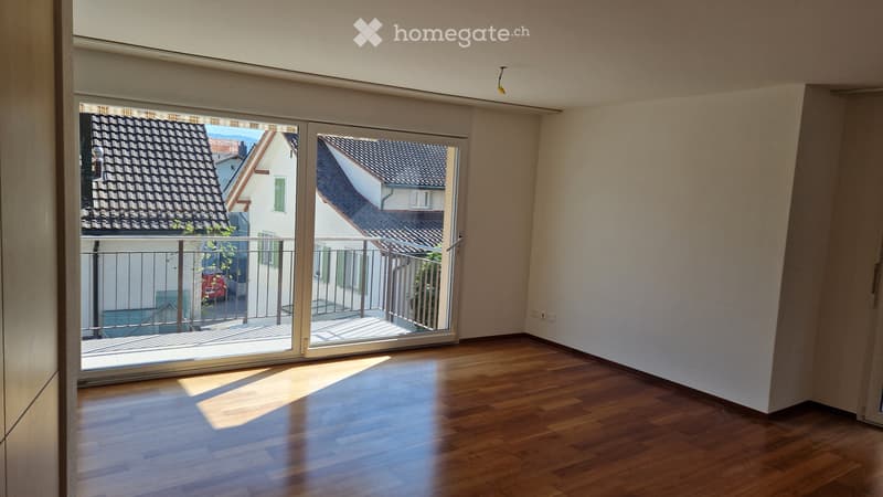 Empty room with wooden ceiling, large window offering a view of neighboring rooftops and a balcony area.