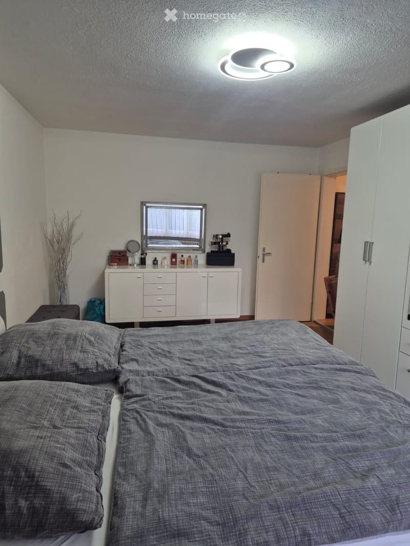 Modern bedroom with a large bed, grey bedding, a white cabinet with drawers, a mirror, and a small stool. A vase with dried branches is placed on the floor near the bed.