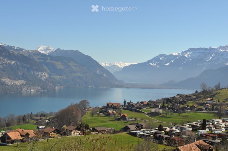 A picturesque village nestled in a lush green valley, surrounded by snow-capped mountains and a serene lake. Traditional houses with red-tiled roofs dot the landscape, creating a tranquil and idyllic setting.