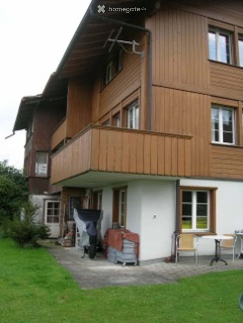 Two-story wooden building with a balcony, white walls, and multiple windows. A small patio area with a table and chairs is visible in front.