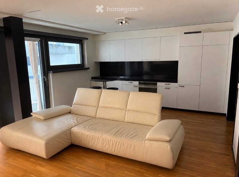Modern, open-plan living and dining area with a white leather sofa, a small dining table with stools, and a sleek kitchen area featuring white cabinets and a black countertop.