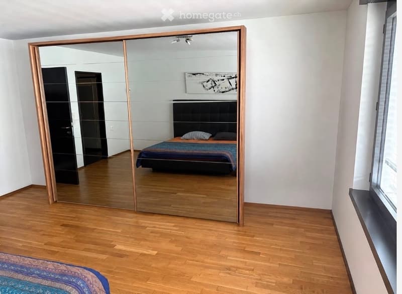 Modern bedroom with wooden flooring, large mirrored wardrobe, bed with colorful bedding, and abstract artwork on the wall.