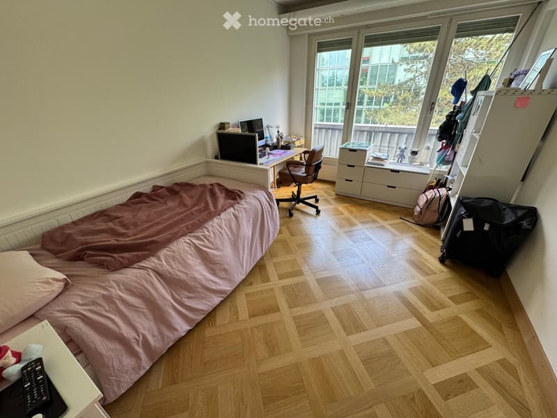 A small, well-lit bedroom with wooden flooring, a single bed with pink bedding, a desk with a laptop and chair, and a storage unit near the window. The room has large windows with a view of the outside.