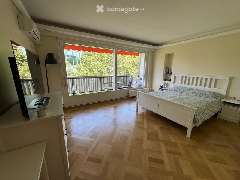 Bright bedroom with wooden flooring, large window opening to a balcony, bed with patterned bedding, nightstand, small table, and lamp.