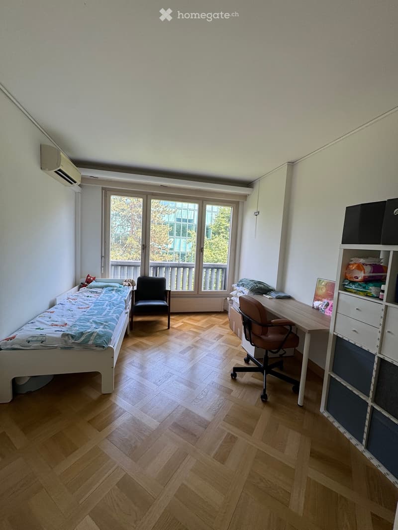 Cozy bedroom with wooden flooring, a single bed with colorful bedding, a desk with a chair, and a shelf with storage boxes. Large windows offer a view of greenery outside.