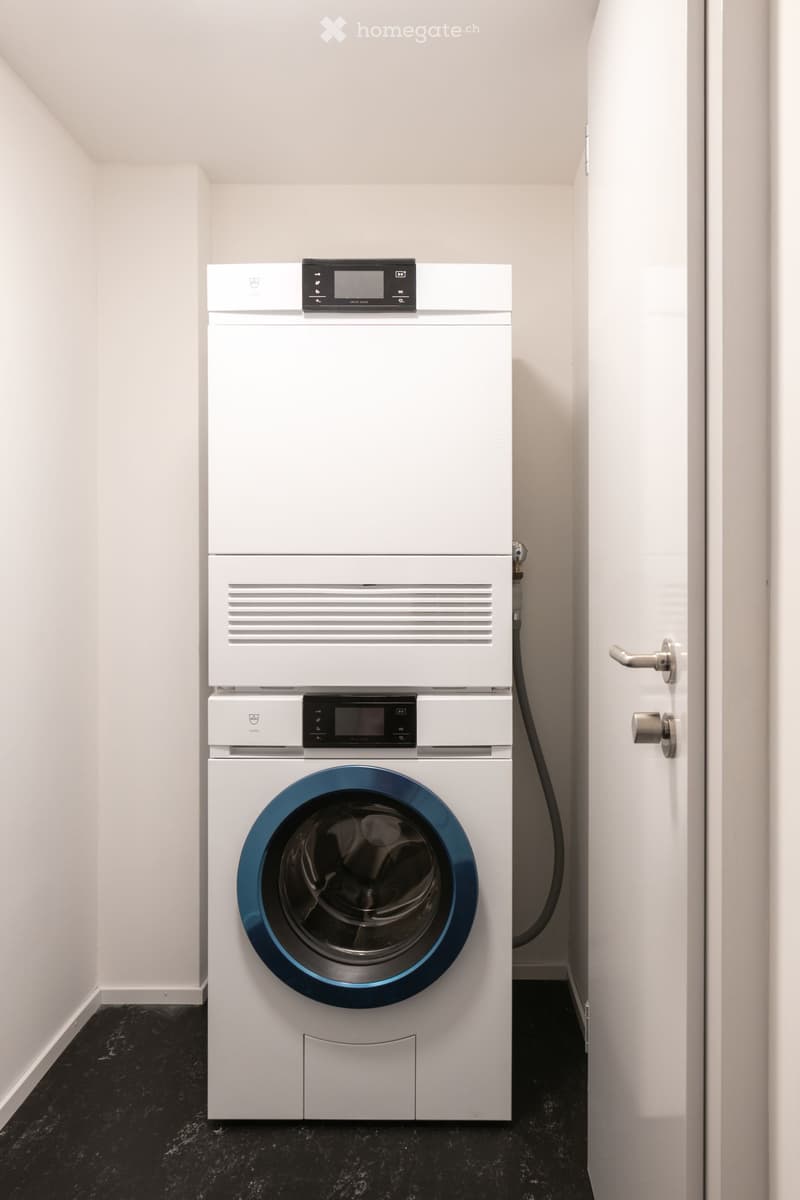 A small, modern laundry room with a white washing machine and dryer stacked vertically, featuring a sleek design and digital control panels.