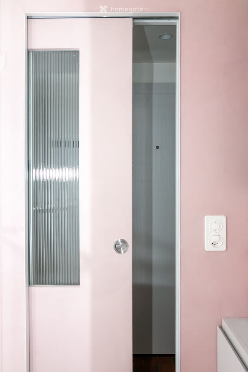 A narrow hallway with light pink walls, a frosted glass window on the left, and a white electrical outlet on the right. The hallway leads to another room with a visible ceiling light fixture.