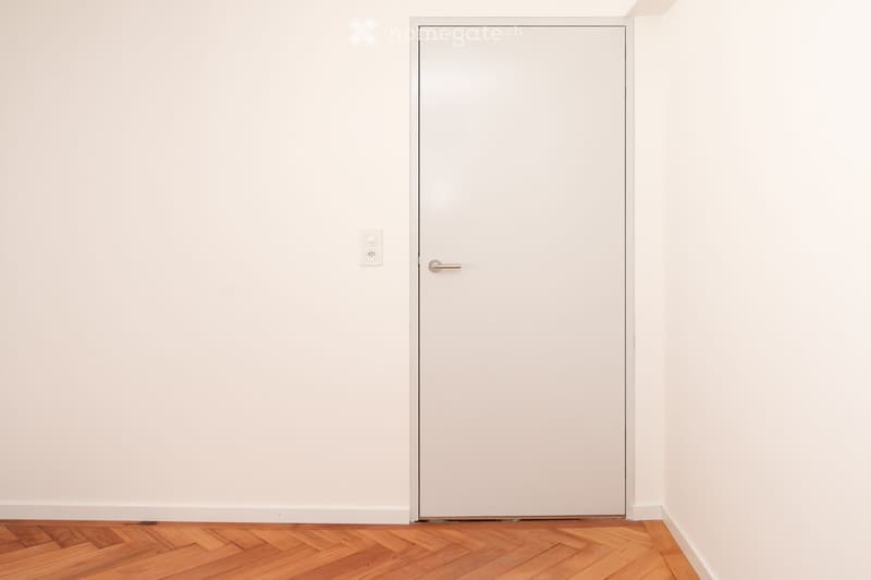 Empty room with white walls, wooden herringbone floor, and a closed white door with a silver handle.