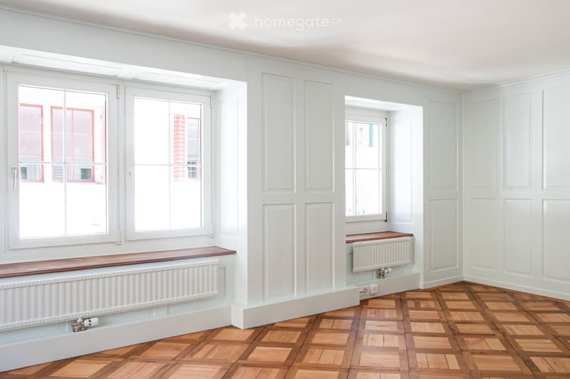 Empty room with light-colored walls, wooden floor, large windows, and built-in wooden benches with radiators underneath.
