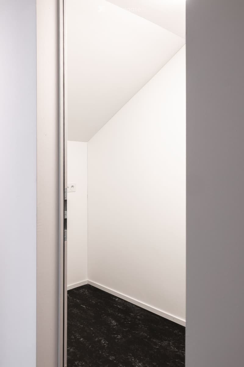 A narrow, minimalist hallway with white walls, a black floor, and a partially open door leading to another room.