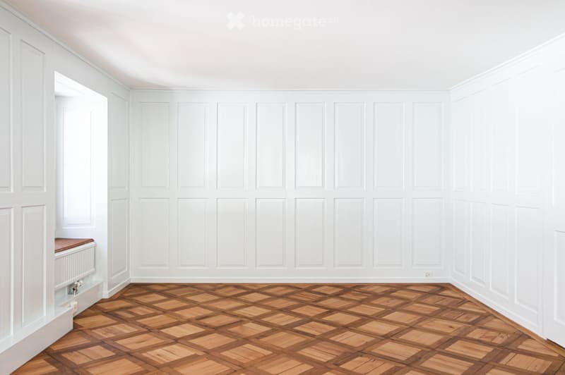 Empty room with white paneled walls and a herringbone wooden floor. No furniture or equipment present.
