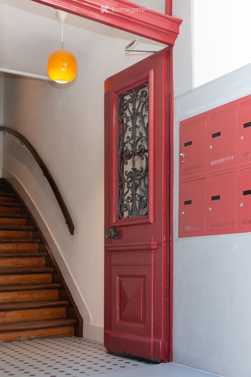 Hallway with a red door featuring ornate metalwork, wooden staircase, and red mailboxes on the wall.