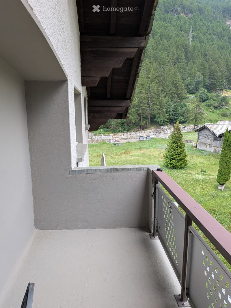 Balcony with wooden staircase leading up, metal railing with decorative pattern, overlooking a green landscape with trees and a small wooden structure.