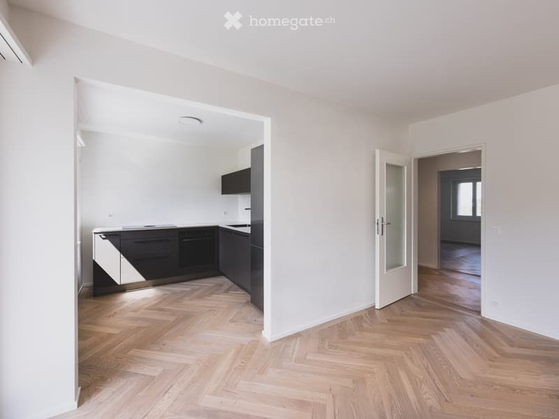 Bright, unfurnished room with wooden herringbone flooring, white walls, and a modern kitchen area visible through an open doorway. The kitchen features sleek black cabinets and stainless steel appliances.