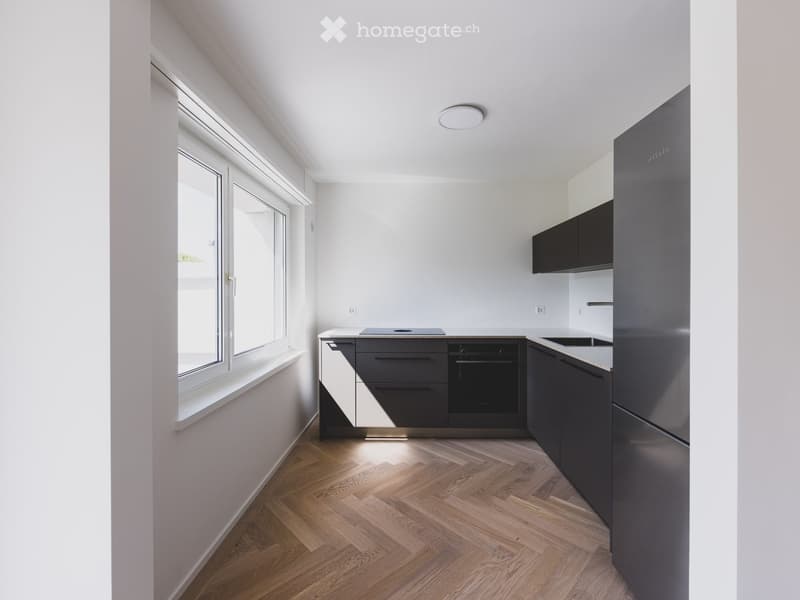 Modern kitchen with wooden flooring, large windows, stainless steel appliances, and dark grey cabinets.