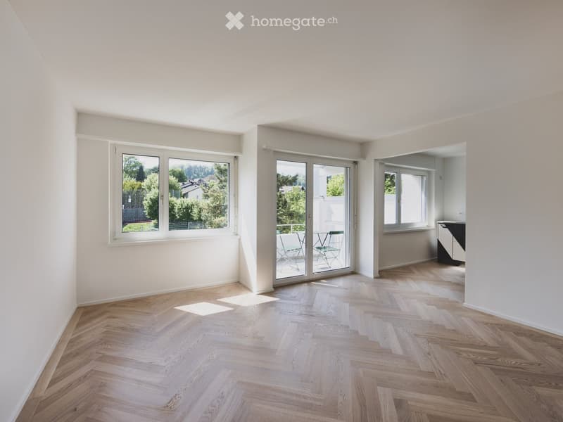 Bright, empty room with wooden herringbone flooring, large windows, and sliding glass doors leading to a balcony area with outdoor furniture.