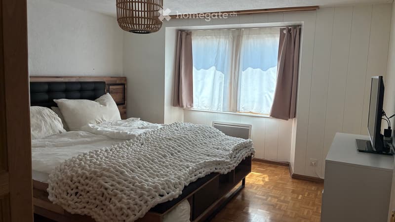 Cozy bedroom with a wooden bed, white bedding, a large window with curtains, and a wicker pendant light.