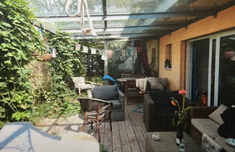 A covered outdoor patio with wooden flooring, featuring a sofa, chairs, a small table, and a flower pot. The space is surrounded by greenery and has a glass roof.