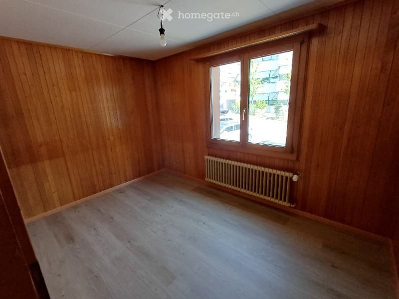 Empty room with wooden walls and floor, a single window, a radiator, and a ceiling light fixture.