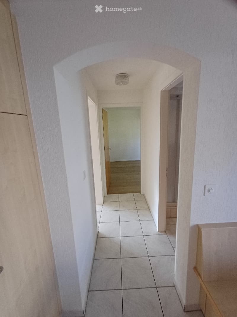A narrow, arched hallway with tiled flooring and white walls, leading to an open doorway at the end.