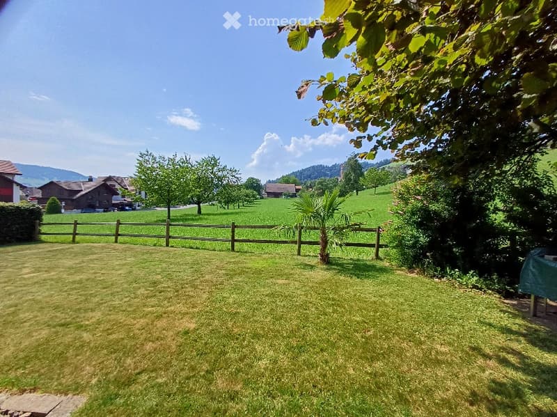 A well-maintained lawn with a wooden fence, surrounded by trees and distant houses in a rural setting.