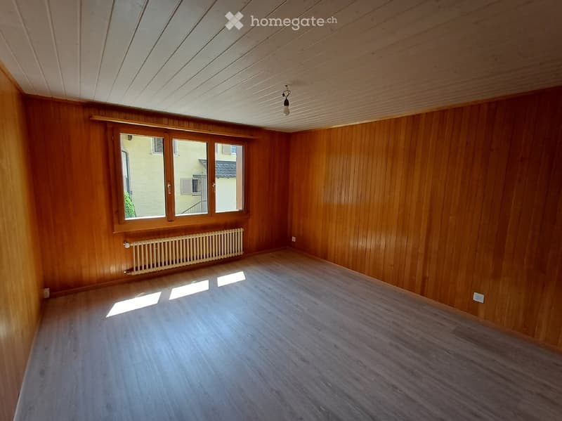 Empty room with wooden walls, wooden floor, large window, radiator, and exposed wooden ceiling beams.