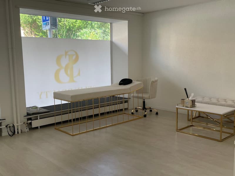 Modern, well-lit room with a massage table, a white chair, a small table with a stool, and a large window with a view of greenery.