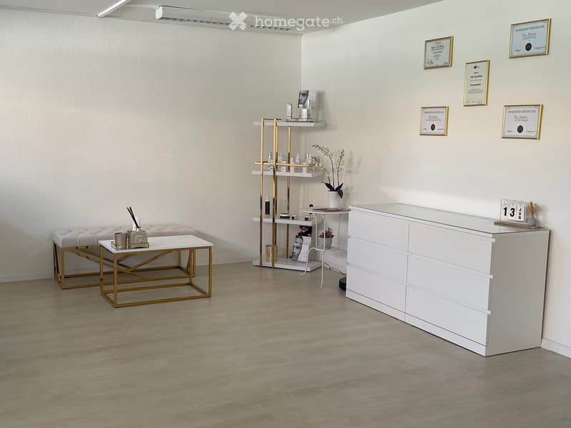 Modern reception area with a white reception desk, small table with decorative items, shelves with bottles and a vase, and framed certificates on the wall.