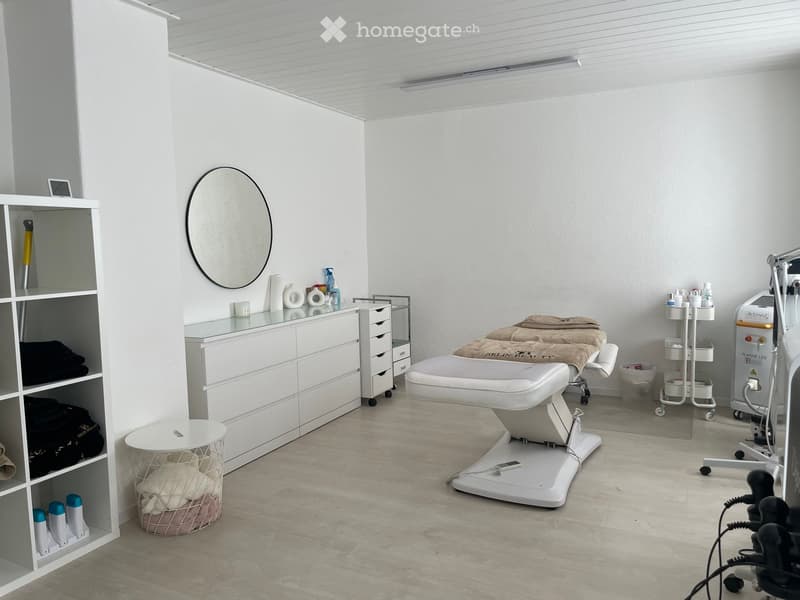 A clean, modern beauty treatment room with a large round mirror, a white treatment table, a rolling cart with supplies, a shelf with towels and products, and a glass-fronted cabinet.