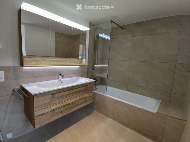 Modern bathroom with a double sink, wooden vanity, large mirror with LED lighting, and a bathtub with a glass shower enclosure.