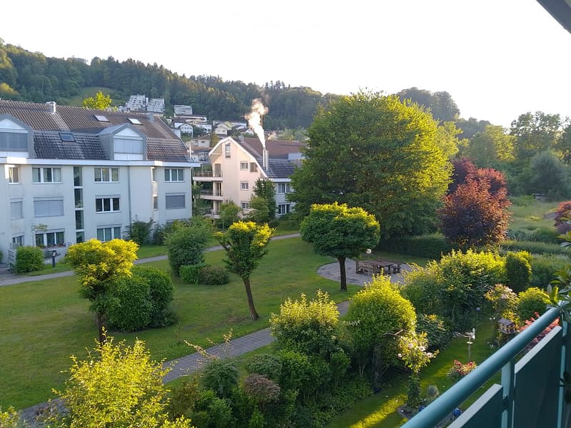 View from a balcony overlooking a well-maintained garden area with pathways, benches, and surrounding residential buildings.