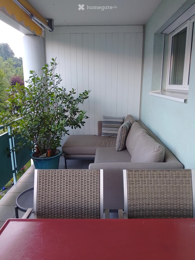 A small, modern balcony with a light blue floor, a white wooden wall, a grey sofa with striped cushions, and a wicker basket. A potted plant adds greenery.
