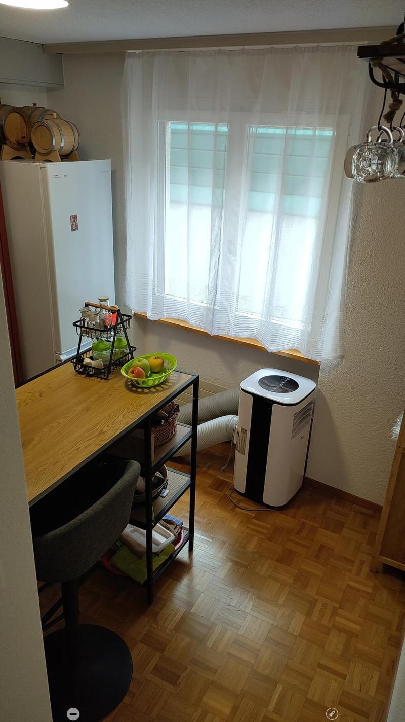 A small, well-lit room with wooden flooring, a table with a fruit bowl, a chair, a refrigerator, and an air conditioner. Wine barrels are stacked against the wall, and a cup rack holds various items.