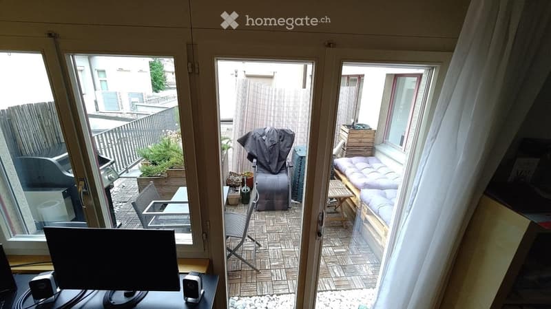 A modern living room with large windows and a sliding glass door leading to a small outdoor patio. The room features a sofa, a chair, a small table, and a laptop on a desk. The patio includes a cushioned bench and some potted plants.