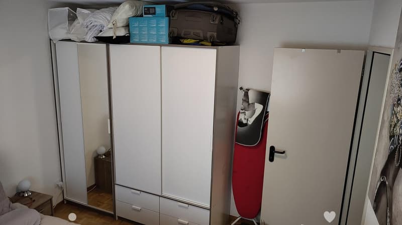 A furnished room with a large white wardrobe, a small nightstand with a lamp, and an ironing board leaning against the wall. The room has wooden flooring and a bed partially visible on the left side.