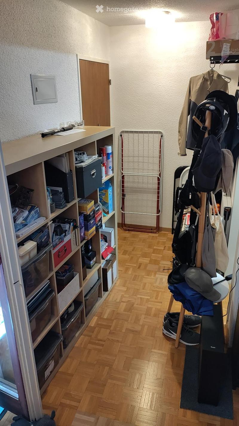 A narrow hallway with wooden flooring, a built-in shelving unit on the left, and a coat rack on the right holding jackets, hats, and bags. A folding chair and a small table are also visible.