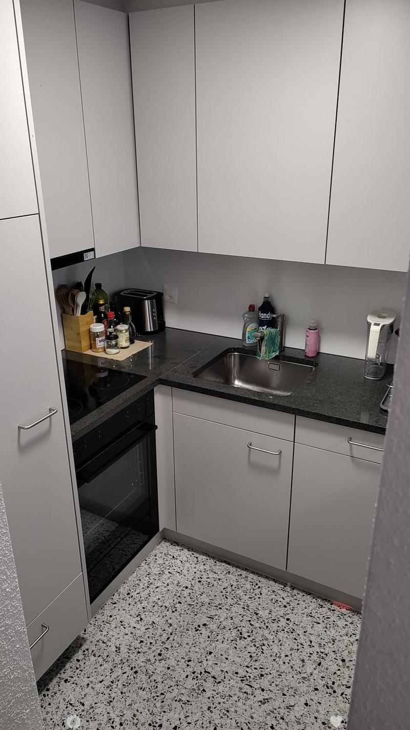 Modern kitchen with white cabinets, black countertop, stainless steel sink, and various appliances including a toaster and oven. The floor has a speckled black and white tile pattern.