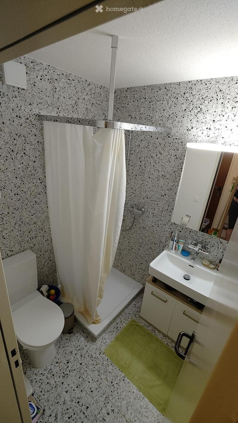 Modern bathroom with white tiles, a toilet, a sink with a mirror, a shower area with a curtain, and a green bath mat.