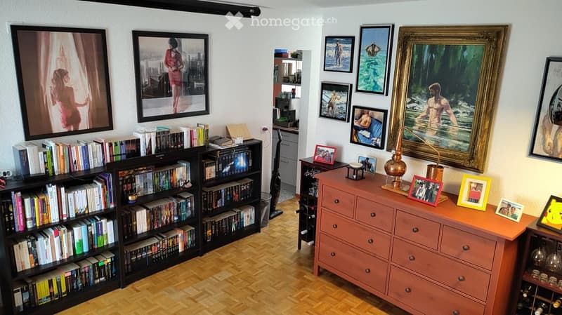 A cozy, well-furnished room with a large bookshelf filled with books, a wooden table with a lamp and picture frames, and a wine rack in the corner.