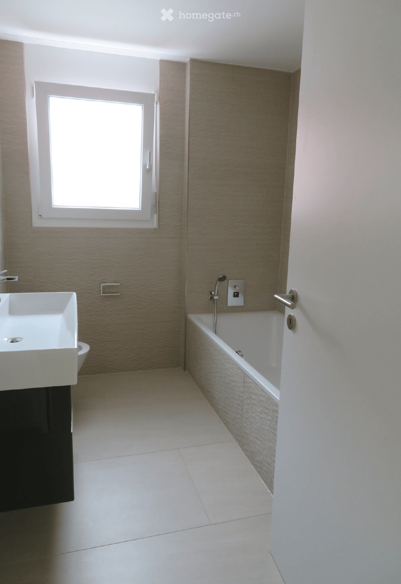 Modern bathroom with white tiles, a white sink, and a bathtub, featuring a window and sleek fixtures.
