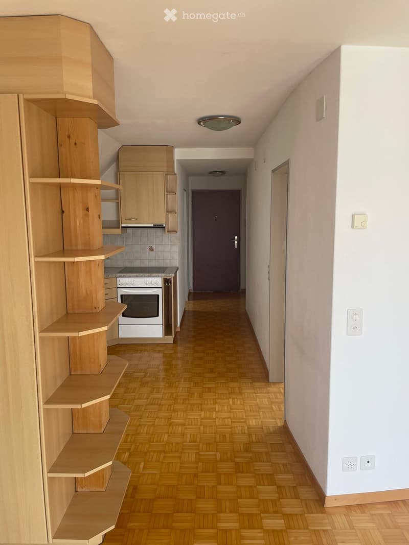 A narrow hallway with wooden flooring, white walls, and a wooden shelving unit on the left side. A kitchen area is visible at the end of the hallway, featuring a white oven, wooden cabinets, and a tiled backsplash.