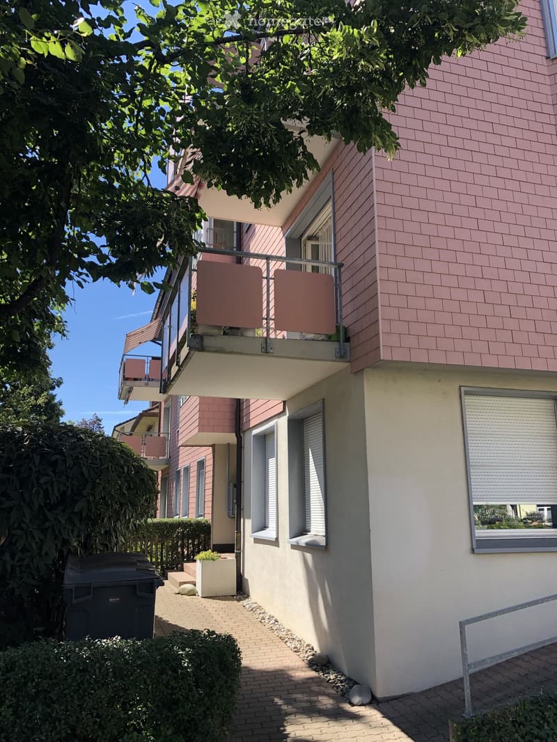 Two-story residential building with pink siding, multiple windows, and balconies. The ground floor has a small garden area with a trash bin and a potted plant.