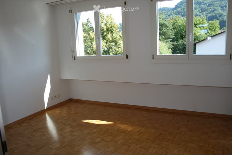 Empty room with wooden flooring and large windows offering a scenic view of trees and mountains.